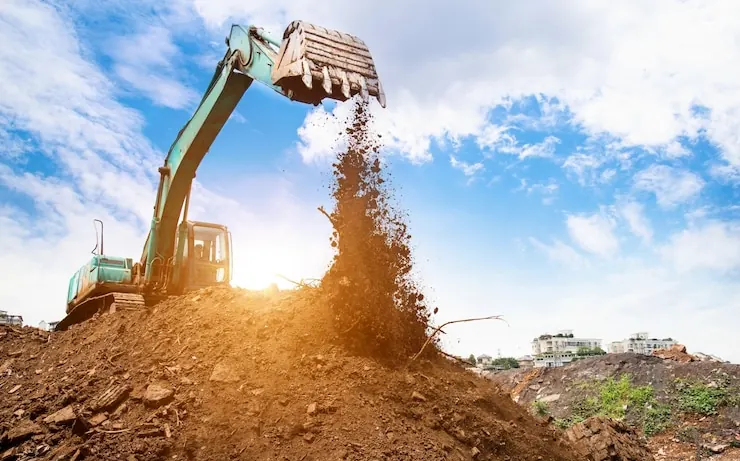 Excavator tipping soil from its bucket onto a mound, houses on the hill behind